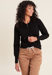 Schwarze Kapuzenjacke mit Reißverschluss und Kordelzug, mit langen Ärmeln und seitlichen Streifen, kombiniert mit beigen Schrumpfjeans mit elastischem Bund und Taschen.