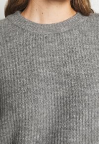 Gros plan sur une personne portant un pull gris côtelé doux et texturé avec un col rond, les longs cheveux bruns visibles.