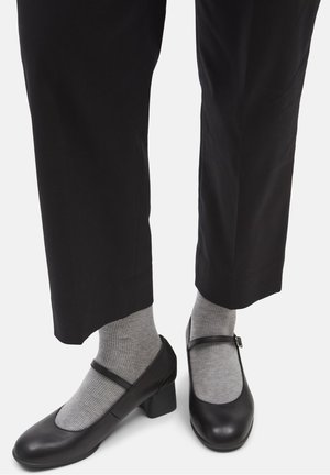 Zapatos Mary Jane de cuero negro con puntera redonda, tacón cuadrado y correa ajustable, combinados con calcetas grises acanaladas. Se ven pantalones negros.