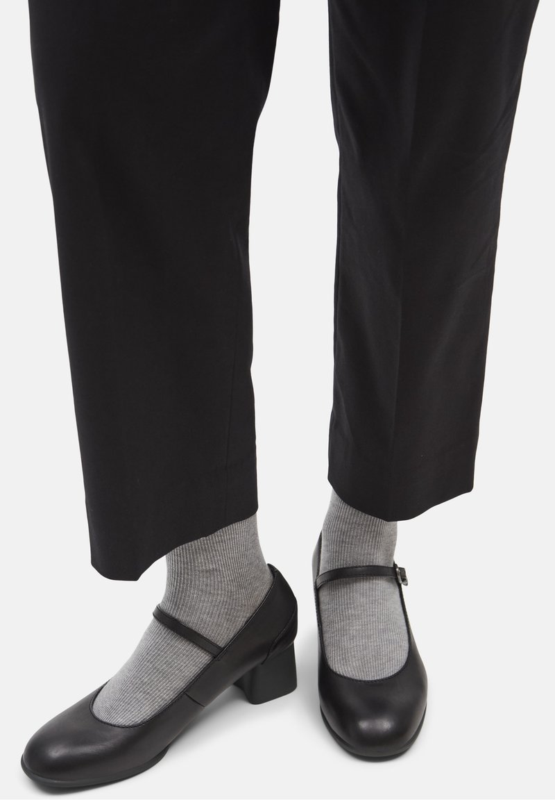 Chaussures Mary Jane en cuir noir avec bout arrondi, talon bloc et bride ajustable, portées avec des chaussettes grises côtelées. Pantalon noir visible.