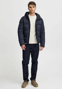 Pufferjakke i marineblå med hætte, har et quiltet design og trykknaplukninger; parret med en cremefarvet sweater, mørke jeans og grå sko.
