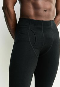 Čierne legíny z pružného materiálu s predným otvorom, hladkou textúrou a kontrastným šitím pozdĺž švov.