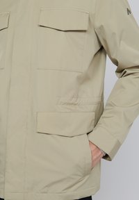 Beige jakk, mis on valmistatud pehmest materjalist, kahte rinnaõmblust, küljetaskut ja elastseid mansette. Minimalistlik disain puhaste joontega.