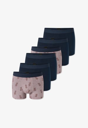 Jeune homme musclé souriant, portant un boxer mauve avec un imprimé ananas foncé et une ceinture noire, debout devant un fond uni.