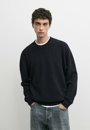 Jeune homme aux cheveux bruns bouclés, portant un pull noir et un jean bleu, debout les mains dans les poches devant un fond clair uni.