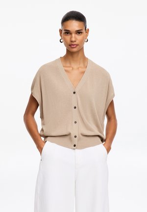 Femme portant un cardigan beige à manches courtes boutonné et un pantalon blanc taille haute à jambes larges, debout, les mains dans les poches.