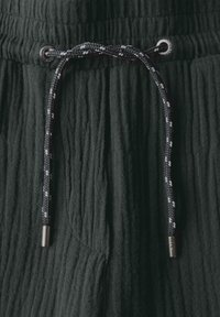 Tmavě zelená textura látky se zvrásněným vzorem, obsahující černobílou spletenou tkaničku s kovovými prvky.