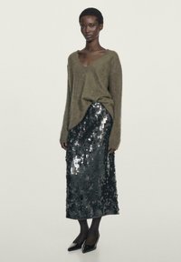 Woman wearing an olive green V-neck sweater, black sequined midi skirt, black tights, and black pointed-toe heels standing against a plain background.