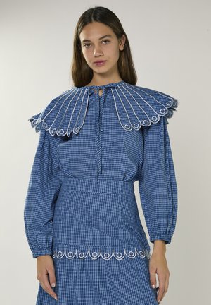 Junge Frau trägt ein blau kariertes Kleid mit großem plissiertem Kragen und weißen bestickten kreisförmigen Mustern am Kragen und an der Taille.