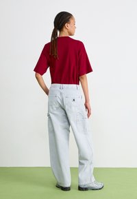 Maglietta rossa a maniche corte e pantaloni cargo grigio chiaro con diverse tasche. La donna indossa scarpe da ginnastica grigie. Lo sfondo è semplice.