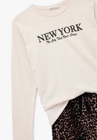 Lichtbeige langarmshirt met zwarte tekst "NEW YORK" en "The City That Never Sleeps"; gecombineerd met zwarte en bruine luipaardprint broek.