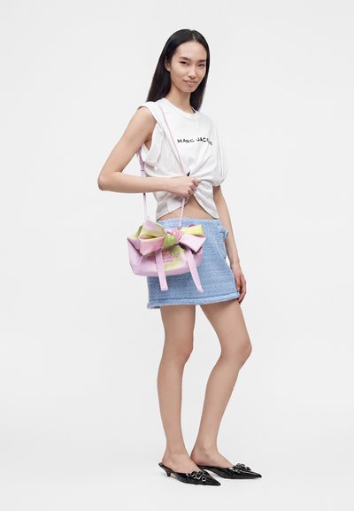 Γυναίκα φοράει λευκό αμάνικο τοπ Marc Jacobs, μπλε μίνι φούστα, μαύρα μυτερά παπούτσια και κρατάει ένα ροζ τσαντάκι με μεγάλο φιόγκο.