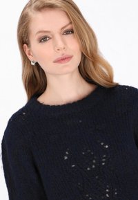 Granatowy sweter dzianinowy o luźnym kroju, z teksturowanym wzorem i szerokim dekoltem. Modelka nosi srebrne prostokątne kolczyki.