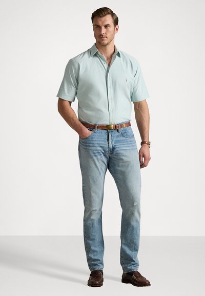 Lysegrønn kortermet skjorte, tettsittende, med et lite emblem; parret med lyseblå jeans med slitte detaljer og brun belte.