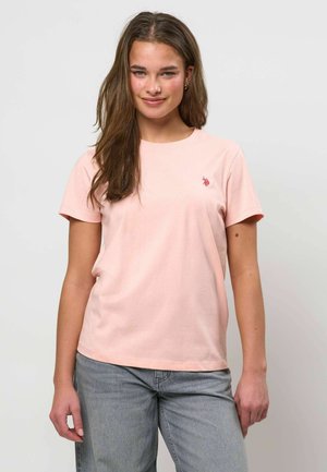 Mladá žena s dlhými hnedými vlasmi, oblečená v svetloružovom krátkom tričku a svetlomodrých džínsoch, stojaca pred jednoduchým pozadím.