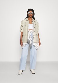 Casaco oversized bege claro, top branco cropped, fita de atar em volta da cintura em tie-dye, calças largas de ganga azul claro e botins brancos.