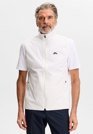 Starszy mężczyzna z siwymi włosami i brodą, ubrany w białą kamizelkę na zamek błyskawiczny, noszącą pod nią krótkie rękawy koszuli, stoi na jednolitym jasnym tle.