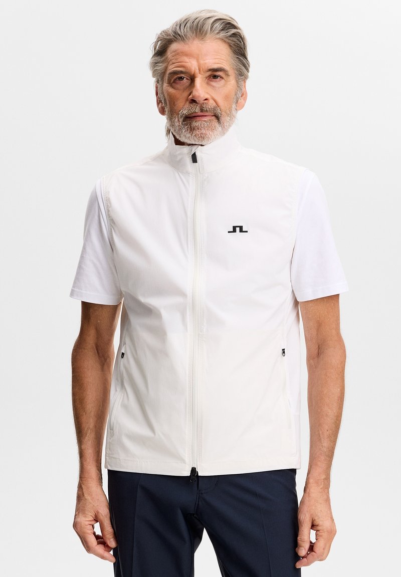 Oudere man met grijs haar en baard, gekleed in een witte vest met rits over een shirt met korte mouwen, staand tegen een effen lichte achtergrond.