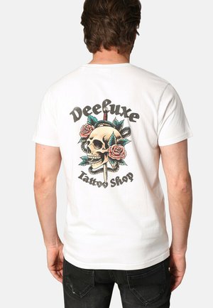 T-shirt en coton blanc arborant un grand graphisme d'un crâne avec des roses et une dague, ainsi que le texte "Deebuxe Tattoo Shop" en lettres gras.