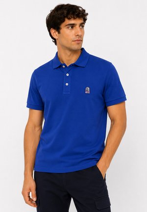 Homme portant un polo bleu avec un logo sur la poitrine et un pantalon cargo noir, debout avec une main dans la poche, regardant sur le côté.