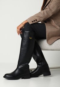 Černé kožené boty po kolena s hladkým povrchem, plochou podrážkou a zlatými akcenty. Nošeny s černými kalhotami a kostkovaným blejzrem.