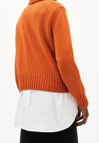 Personne portant un pull en maille orange sur une chemise blanche avec un pantalon noir, vue de dos sur un fond blanc uni.