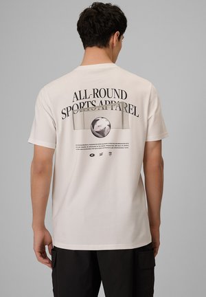 Mężczyzna w białej koszulce z czarnym napisem "All-Round Sports Apparel" i grafiką piłki nożnej na plecach, stojący na szarym tle.