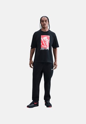 Mand med fletninger iført en sort t-shirt med rød og hvid grafik, sorte bukser og sorte sneakers med røde accenter.
