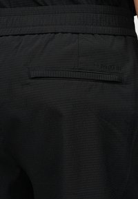 Κοντινή λήψη πίσω τσέπης μαύρου υφασμάτινου παντελονιού με μικρό λογότυπο Calvin Klein κεντημένο πάνω από το άνοιγμα της τσέπης.