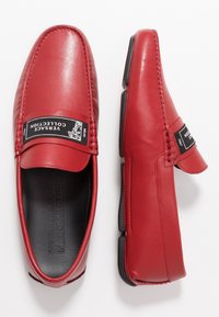 Rote Lederloafers mit flacher Sohle, gestickten Details und einem schwarzen Markenetikett auf der Oberseite. Glatte Oberfläche und elegantes Design.