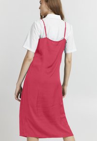 Abito midi fucsia con spalline sottili e taglio dritto. Presenta una texture liscia e un design semplice. Indossato sopra una camicia bianca con colletto.