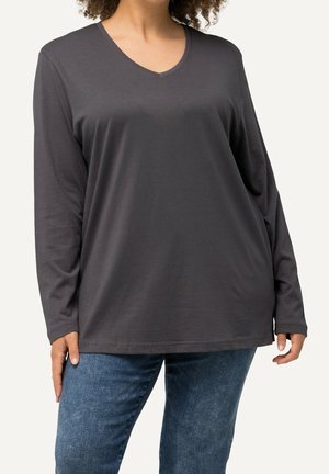 Femme portant un t-shirt ample gris foncé à manches longues et col en V, et un jean bleu, debout devant un fond clair uni.
