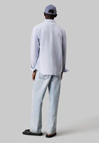 Camicia a maniche lunghe di un blu chiaro con colletto a botton-down, abbinata a jeans larghi in lavaggio chiaro e sandali neri. Un berretto grigio completa il look.