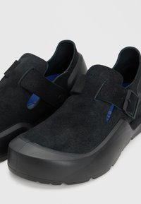 Ζεύγος μαύρων slip-on παπουτσιών με επάνω μέρος από σουέτ, φαρδύ ρυθμιζόμενο λουράκι και παχιά λαστιχένια σόλα σε ανοιχτό γκρι φόντο.