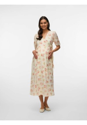 Femme enceinte souriante, portant une robe midi légère à motifs floraux avec des manches bouffantes et des ballerines beiges, debout devant un fond blanc uni.