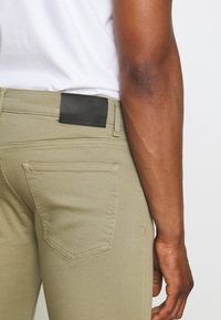 Beige dressade byxor i texturerat tyg, en svart läderlapp på midjan och standard bakfickor synliga.