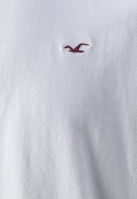 Balta kokvilnas T-kreklu ar teksturētu virsmu, ar mazu bordo izšūtu logotipu ar lidotāju putnu kreisajā augšējā stūrī.