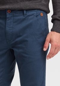 Pantalons en coton bleu foncé avec une texture lisse, dotés d'un bouton marron et d'une petite étiquette en cuir sur la poche avant.