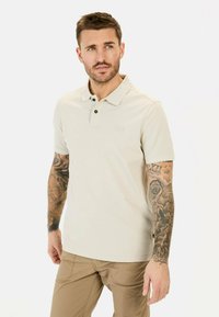 Beige polo med krave, to-knaps placket og diskret logodetalje. Modellen har tatoverede arme og ifører sig brune bukser.