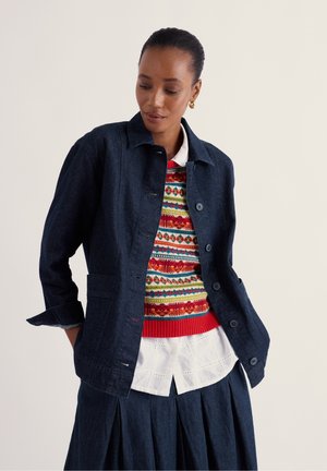 Femme portant une veste en jean foncée par-dessus un pull coloré à motifs, une chemise blanche à broderies anglaises et une jupe plissée sombre sur un fond clair uni.