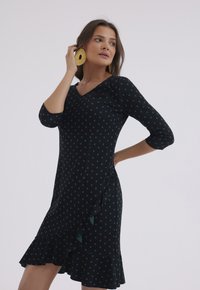 Czarna sukienka w turkusowe kropki, z dekoltem w kształcie V, z rękawami trzeci długości i falbaną u dołu. Miękka tkanina z dopasowanym krojem.