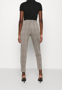 Kostkované kalhoty v barvě světle hnědé a černé, na míru střihané s vysokým pasem a úzkými nohavicemi, vyrobené z měkké textilie. Nošeny s černými sandály na vysokém podpatku.