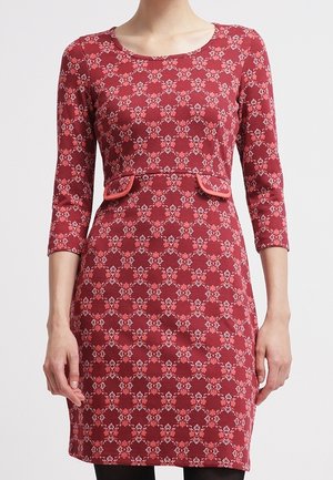 Woman wearing a fitted knee-length red dress with a geometric floral pattern, 3/4 sleeves, and decorative flap pockets at the waist.