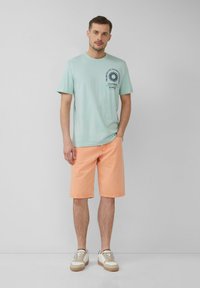 Lichtgroene katoenen t-shirt met een marineblauw grafisch ontwerp op de borst, gecombineerd met perzikkleurige denimshorts en witte sneakers met tan accenten.