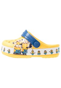 Gula träskor med blå rem, prydda med Minion-karaktärsgrafik på sidan. Tillverkade av lätt material med ventilationshål.