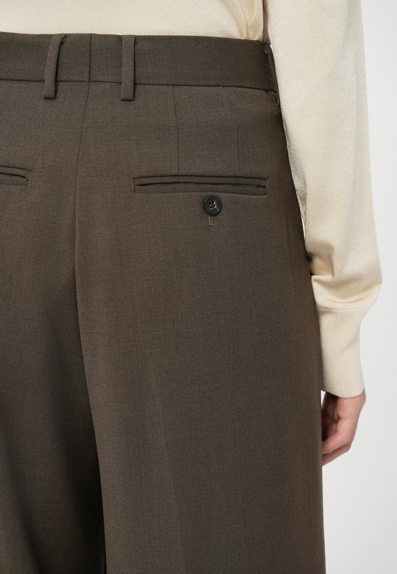 Brown tailored trousers with a smooth texture, featuring two back pockets and a button closure. Paired with a cream-colored sweater.