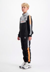 Zwart en grijs trainingspak met oranje accenten, bestaande uit een top met kwart rits en een broek met elastische tailleband, gecombineerd met witte sneakers.
