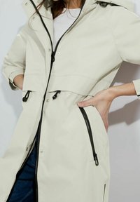 Svetle béžová vodotěsná bunda se zipem vpředu, nastavitelnou kapucí a elastickými manžetami; obsahuje dvě černé boční kapsy na zip.