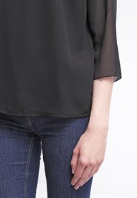 Czarna, przezroczysta bluzka z trzyczwartowymi rękawami, gładka w dotyku i luźna, połączona z ciemnoniebieskimi rurkami. Widoczna ręka spoczywająca na dżinsach.