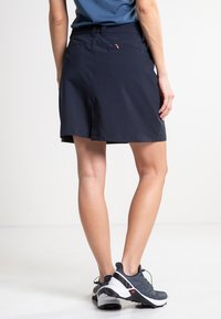 Mörkblå skorts med en mjuk textur, med midja, sidofickor och en lätt A-linjeform. Bärs med gråa träningsskor.